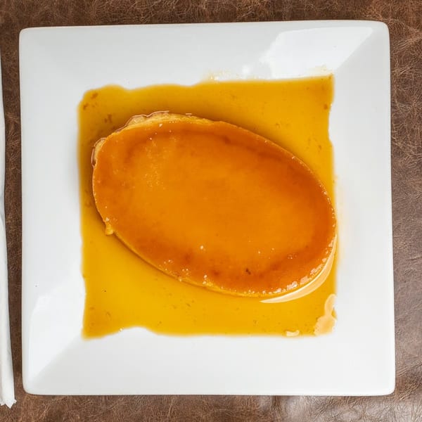 A rectangular white plate holds an oval flan dessert with golden caramel sauce.