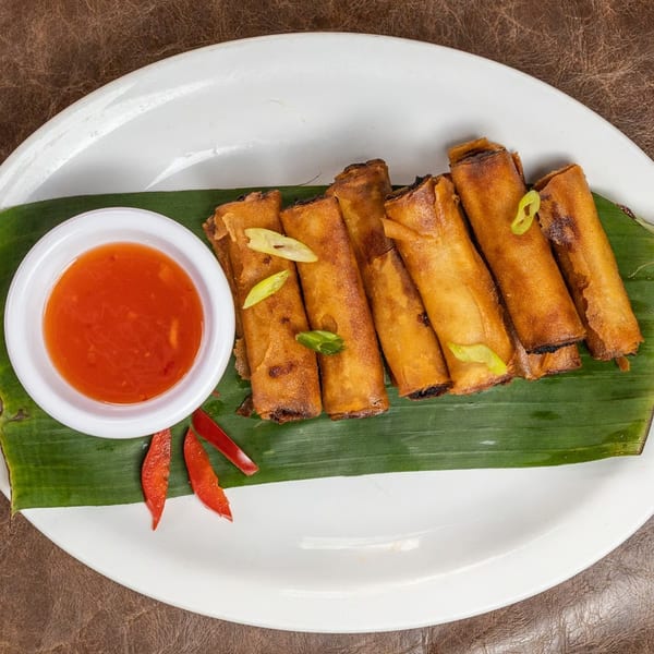 Crispy fried spring rolls with sweet chili dipping sauce and chili slices on a banana leaf.