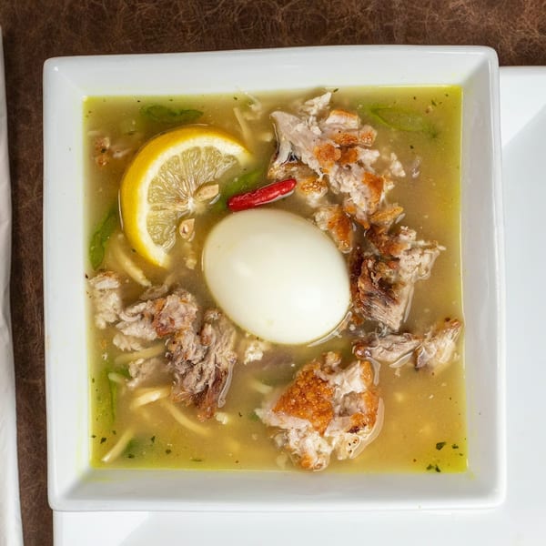 A bowl of Filipino Mami soup with chicken, noodles, a hard-boiled egg, lemon, and chili.